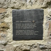 Eine Gedenktafel mit dem Gebet des Vertrauens ist in eine alte, verwitterte Steinwand eingebettet.