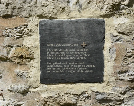Eine Gedenktafel mit dem Gebet des Vertrauens ist in eine alte, verwitterte Steinwand eingebettet.