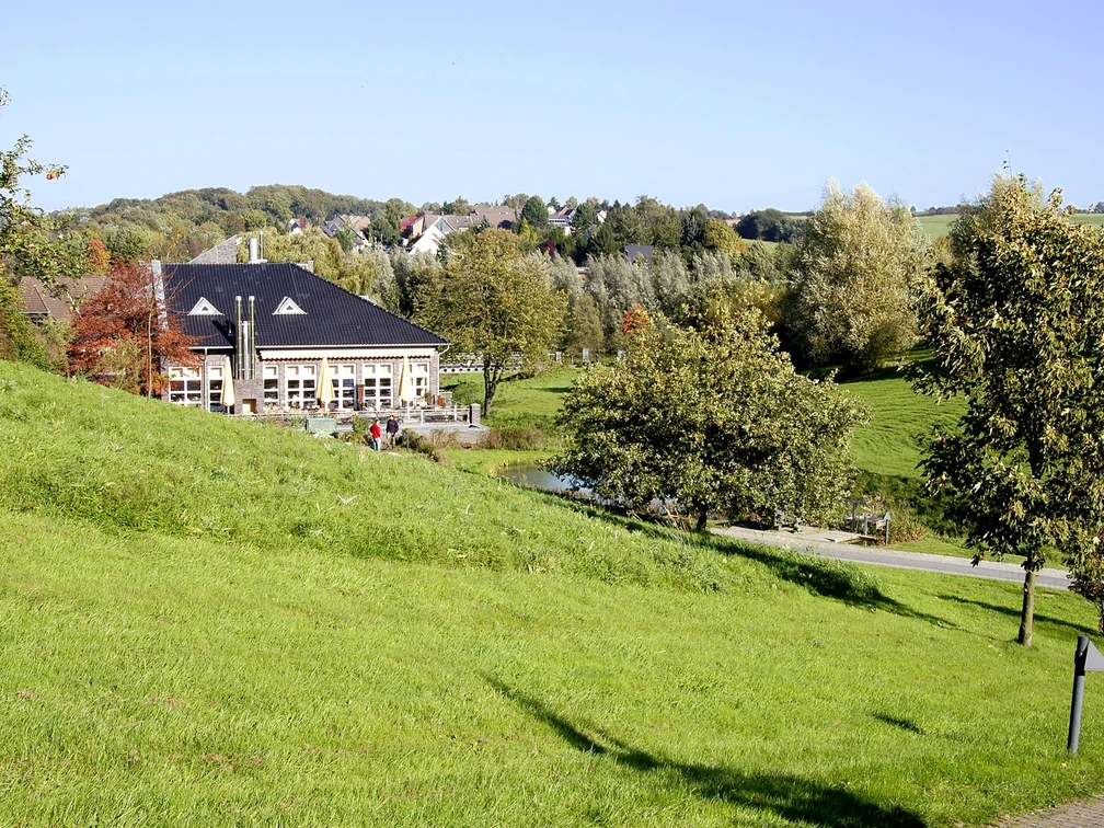 Golf Club Grevenmühle in Ratingen Ein modernes Clubhaus liegt eingebettet in eine weitläufige, grüne Parklandschaft mit vereinzelten Bäumen.