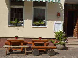 Zwei rustikale Holzbänke und Tische vor einem Gasthofeingang mit Blumenschmuck und Markise.