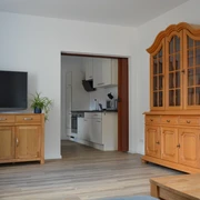 Wohnzimmer mit Holzmöbeln und Blick in eine moderne Küche; heller Holzfußboden, Pflanzen im Raum.