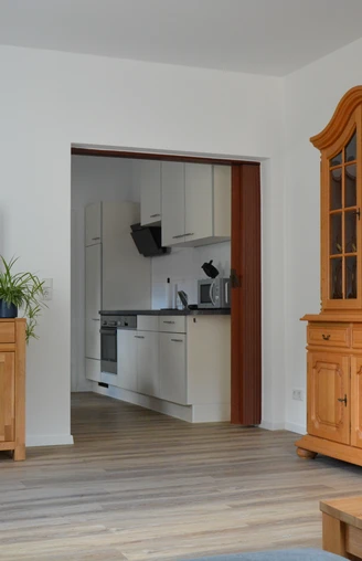 Wohnzimmer mit Holzmöbeln und Blick in eine moderne Küche; heller Holzfußboden, Pflanzen im Raum.