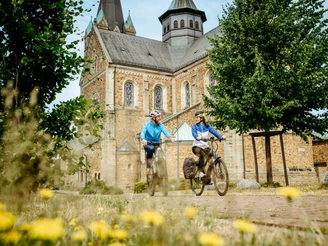 Radfahren auf der Artland-Rad-Tour Ein Paar radelt in sportlicher Kleidung neben einer historischen Backsteinkirche im Sommer.