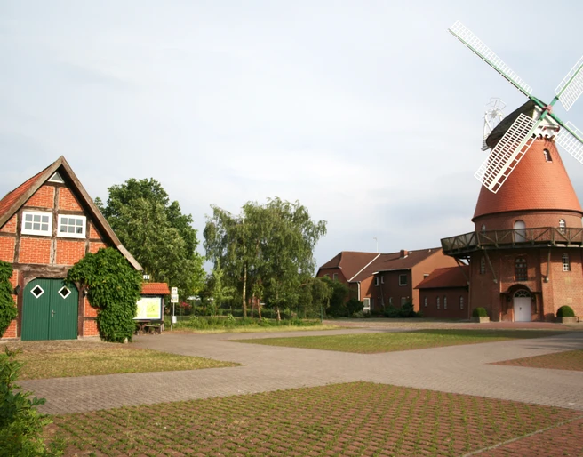 Landesbergen Hochzeitsmühle Rote Backsteinmühle mit weißen Flügeln neben grünem Fachwerkhaus, umgeben von Bäumen und Weg.