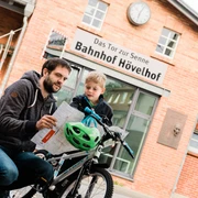 Mann und Kind betrachten eine Karte vor dem Backsteingebäude des Bahnhofs Hövelhof.