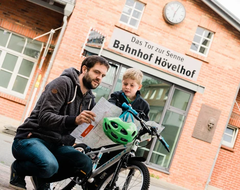 Mann und Kind betrachten eine Karte vor dem Backsteingebäude des Bahnhofs Hövelhof.