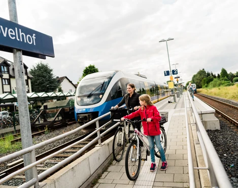 Zwei Menschen schieben im Bahnhof Hövelhof Fahrräder am Bahnsteig entlang, rechts ein vorbeifahrender Zug.