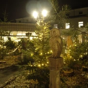 Weihnachtsmarkt-in-Heiligenhaus-Eulenskulptur.JPG Eine Holzskulptur einer Eule steht zwischen beleuchteten Tannenbäumen auf einem Weihnachtsmarkt bei Nacht.