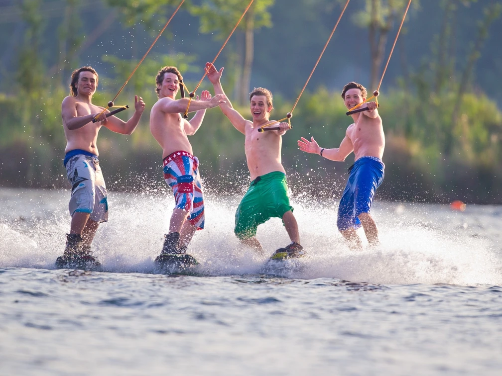 Wasserskianlage Langenfeld Vier junge Männer stehen auf Wakeboards, lachen und posieren fröhlich auf einer Wasserskianlage.
