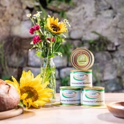 Gut Schobbenhaus aus Mettmann Verschiedene Dosen Wurst und frische Sonnenblumen auf einem rustikalen Holztisch vor einer Steinwand.