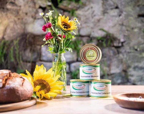 Gut Schobbenhaus aus Mettmann Verschiedene Dosen Wurst und frische Sonnenblumen auf einem rustikalen Holztisch vor einer Steinwand.