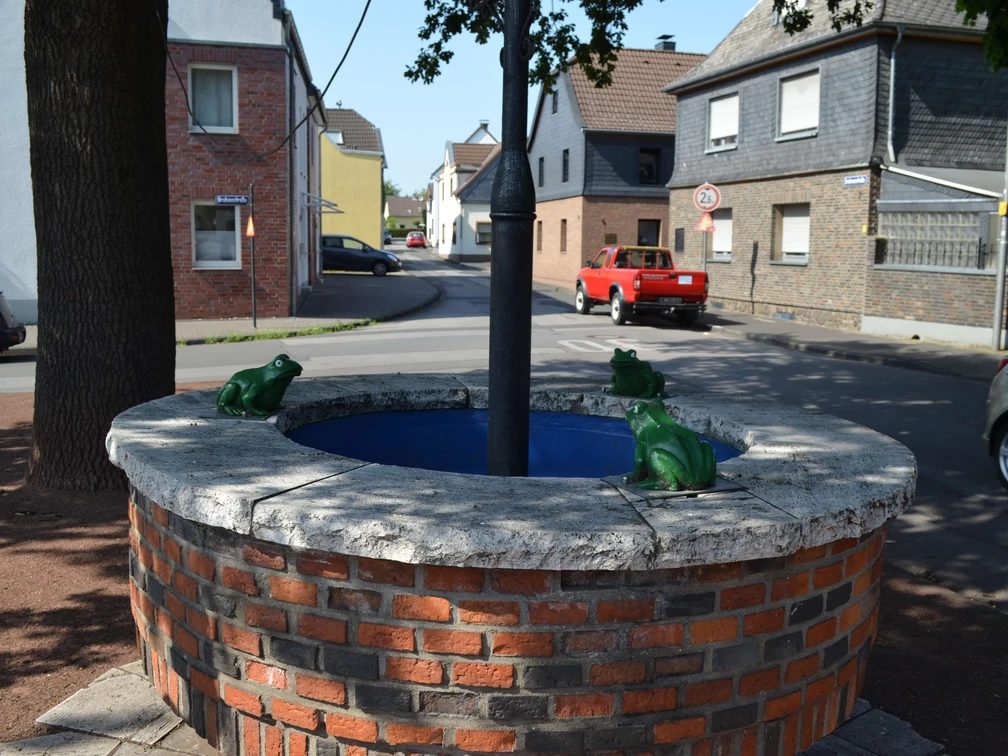 Kradepohl in Monheim am Rhein Ein runder Brunnen aus Ziegelsteinen mit Froschfiguren steht an einer Straßenkreuzung in Monheim am Rhein.