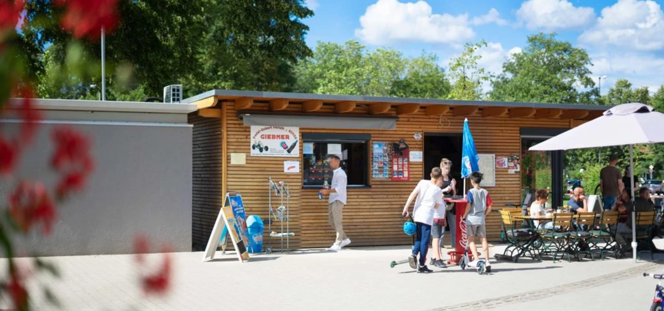 Tourist-Information Giebner in Monheim am Rhein Tourist-Information mit Holzverkleidung, Menschen und Fahrradführungen, umgeben von grünem Park.