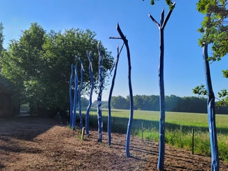 Sieben blau bemalte Holzstämme stehen auf einer Wiese, umgeben von Feldern und Bäumen im Sonnenschein.