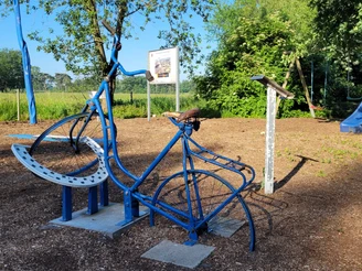 Blaues Fahrraddenkmal im Freien, umgeben von grünen Bäumen, nahe einem Feldweg.