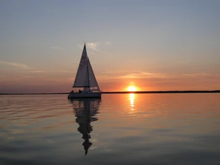 Segeln im Sonnenuntergang Segelboot gleitet ruhig über stilles Wasser, während die strahlende Sonne am Horizont untergeht.
