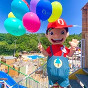 Maskottchen Oskar  Oskarshausen: Ein fröhliches Maskottchen in rotem Outfit hält bunte Luftballons in einem Freizeitpark; im Hintergrund sind Bäume und Spielbereiche zu sehen.