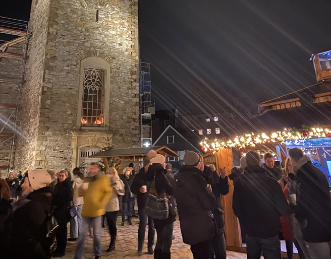 Weihnachtsmarkt2021_Platz Am Offers_StadtVelbert_Schuster_image00036.jpeg Menschen genießen den Weihnachtsmarkt rund um eine beleuchtete Kirche mit Turm im Abendlicht.