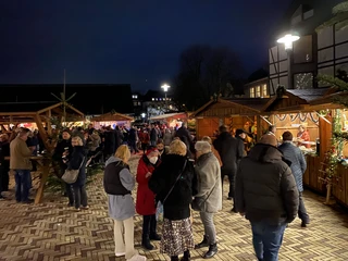 Weihnachtsmarkt2021_Platz Am Offers_StadtVelbert_Schuster_image00048.jpeg Menschen bummeln auf einem abendlichen Weihnachtsmarkt mit festlich geschmückten Holzbuden.