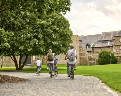 MüGa / Darlington-Park, Mülheim an der Ruhr Vier Radfahrende auf einem Weg im Park, flankiert von Bäumen und historischem Gebäude im Hintergrund.