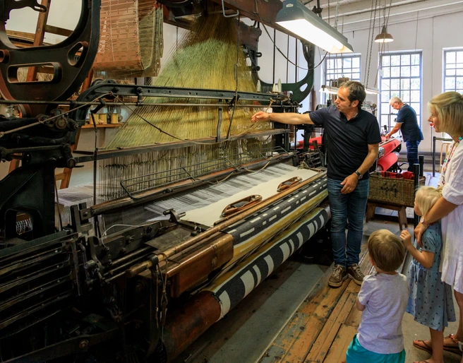 Führung an laufenden Maschinen Historische Webmaschine mit Besuchergruppe, darunter Erwachsene und Kinder, in einem Museum.