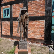 Skulptur "Der Häusling" Holzskulptur eines Mannes mit Stock vor einer Backsteinmauer, flankiert von Fenster und Blumenkasten.