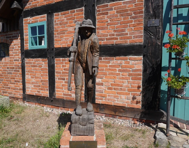 Skulptur "Der Häusling" Holzskulptur eines Mannes mit Stock vor einer Backsteinmauer, flankiert von Fenster und Blumenkasten.