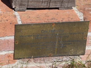 Skulptur "Der Häusling" Die Tafel an einer Backsteinmauer informiert über die Skulptur "Der Häusling" von Ragnar Reusch-Klinkenberg.