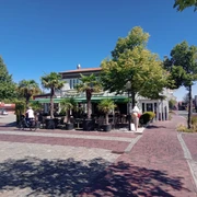 Brasserie am Markt in Apen  Die Brasserie am Markt in Apen präsentiert sich mit einer einladenden Terrasse, gesäumt von Palmen, die unter strahlend blauem Himmel Schatten spenden. Vorne auf dem Platz fährt ein Radfahrer vorbei, während das Gebäude im Hintergrund eine lebendige kulinarische Atmosphäre verspricht.
