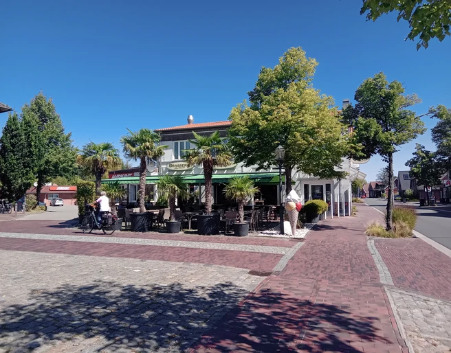 Brasserie am Markt in Apen  Die Brasserie am Markt in Apen präsentiert sich mit einer einladenden Terrasse, gesäumt von Palmen, die unter strahlend blauem Himmel Schatten spenden. Vorne auf dem Platz fährt ein Radfahrer vorbei, während das Gebäude im Hintergrund eine lebendige kulinarische Atmosphäre verspricht.