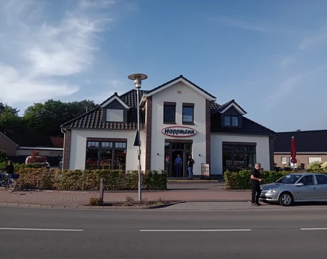 Bäckerei Hoppmann in Augustfehn Das Bild zeigt die Fassade der Bäckerei Hoppmann in Augustfehn. Das Gebäude hat eine traditionelle, einladende Architektur mit einem markanten Schriftzug „Hoppmann“ über dem Eingang. Vor der Bäckerei sind mehrere Personen und geparkte Fahrräder zu sehen.