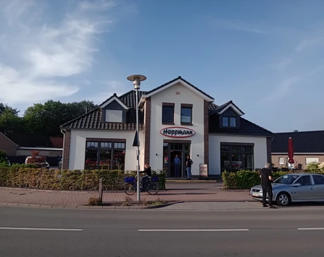 Bäckerei Hoppmann Das Bild zeigt die Fassade der Bäckerei Hoppmann. Das Gebäude hat ein rotes Ziegeldach und weiße Wände, mit einem großen Ladenfenster auf jeder Seite des zentralen Eingangs. Der Schriftzug „Hoppmann“ ist über dem Eingang angebracht. Vor dem Gebäude sind Fahrräder und parkende Autos zu sehen.