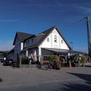 Gaststätte Gleis 3 in Augustfehn Das Bild zeigt die Gaststätte "Gleis 3" in Augustfehn. Das weiße Gebäude im traditionellen Design hebt sich mit einem markanten, dunklen Satteldach hervor. Davor sind Fahrräder und Pflanzen zu sehen, was eine gemütliche, einladende Atmosphäre schafft.