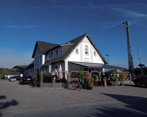 Gaststätte Gleis 3 in Augustfehn Das Bild zeigt die Gaststätte "Gleis 3" in Augustfehn. Das weiße Gebäude im traditionellen Design hebt sich mit einem markanten, dunklen Satteldach hervor. Davor sind Fahrräder und Pflanzen zu sehen, was eine gemütliche, einladende Atmosphäre schafft.