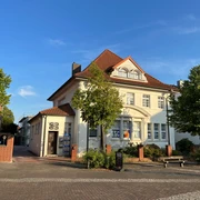 Escape Room Ein großes, weißes Gebäude mit rotem Dach und Escape Room-Schildern an der Fassade, umgeben von Bäumen.