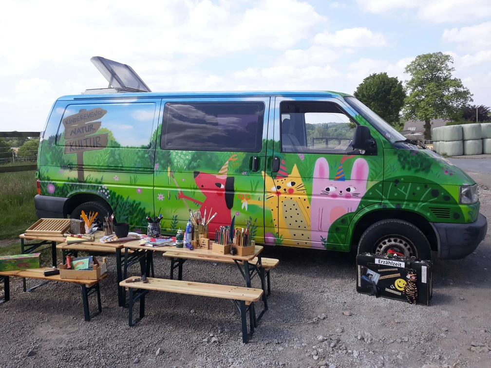 Erzähl- und Papierwerkstatt Mettmann Ein bunt bemalter Van in ländlicher Umgebung, flankiert von Holzbänken und Kunstmaterialien.