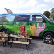 Erzähl- und Papierwerkstatt Mettmann Ein bunt bemalter Van in ländlicher Umgebung, flankiert von Holzbänken und Kunstmaterialien.