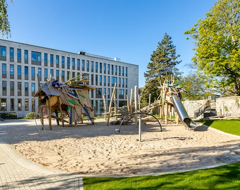 Drachenspielplatz in Ratingen Spielplatz mit Holzstrukturen, Rutsche und Sand, gelegen vor einem modernen Gebäude und umgeben von grünen Bäumen.