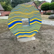 Skulptur Schlussstein Skulptur aus bunten, wellenförmigen Streifen mit zentraler Widmung, umgeben von Backsteinhäusern.