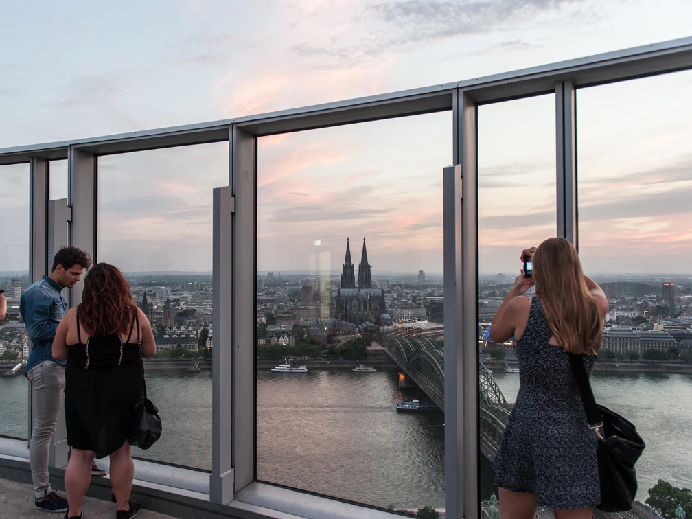 KölnTriangle Personen genießen den atemberaubenden Blick von der Aussichtsplattform des KölnTriangle über die Stadt Köln, die im Abendlicht erstrahlt. Der Panoramablick umfasst den majestätischen Kölner Dom und die glitzernde Rheinlandschaft, was ein unvergessliches Erlebnis bietet.Visitors enjoy the breathtaking view from the viewing platform of the KölnTriangle over the city of Cologne, which shines in the evening light. The panoramic view includes the majestic Cologne Cathedral and the glittering Rhine landscape, offering an unforgettable experience.