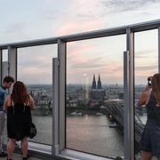 KölnTriangle Personen genießen den atemberaubenden Blick von der Aussichtsplattform des KölnTriangle über die Stadt Köln, die im Abendlicht erstrahlt. Der Panoramablick umfasst den majestätischen Kölner Dom und die glitzernde Rheinlandschaft, was ein unvergessliches Erlebnis bietet.Visitors enjoy the breathtaking view from the viewing platform of the KölnTriangle over the city of Cologne, which shines in the evening light. The panoramic view includes the majestic Cologne Cathedral and the glittering Rhine landscape, offering an unforgettable experience.