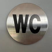 Silbernes, rundes WC-Schild mit schwarzer Aufschrift vor grauem Hintergrund, schlichtes Design.