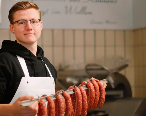 Eigene Wurstherstellung in der Metzgerei Kluke, Mettmann Metzger mit Schürze hält eine Stange frischer Bratwürste in einer traditionellen Metzgerei.