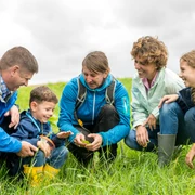 Natur entdecken mit den Naturentdeckern aus Wülfrath Familiengruppe erkundet gemeinsam eine grüne Wiese, alle lächeln und tragen wetterfeste Kleidung.