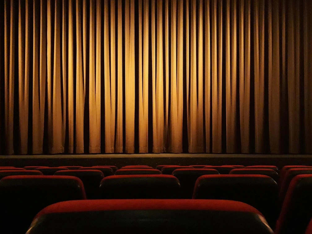 Ein leerer Kinosaal mit roten Sitzen, gerichtete Blicke auf einen geschlossenen roten Vorhang.