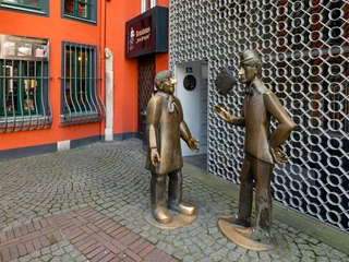 Tünnes and Schäl Monument Die Bronzefiguren von Tünnes und Schäl stehen auf einem gepflasterten Weg vor einer leuchtend roten Fassade. Die Figuren scheinen in ein lebhaftes Gespräch vertieft zu sein. The bronze figures of Tünnes and Schäl stand on a cobbled path in front of a bright red façade. The figures appear to be engrossed in a lively conversation.