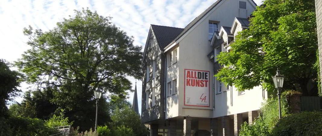 ALLDIEKUNST im Kunsthaus Velbert-Langenberg Ein Kunstzentrum in hellem Gebäude, umgeben von grünen Bäumen und einem klaren blauen Himmel.