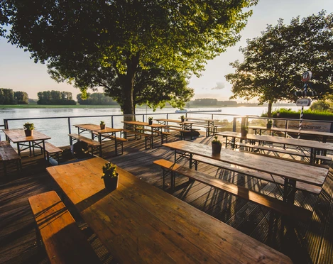 Haus Rheinblick Ott in Monheim Sonnenuntergang über dem Rhein mit leeren Biergartentischen auf einer Holzterrasse im Vordergrund.