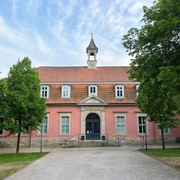 Das Gebäude mit Rosafassade und Glockenturm, umgeben von Bäumen, wirkt ruhig und ansprechend.