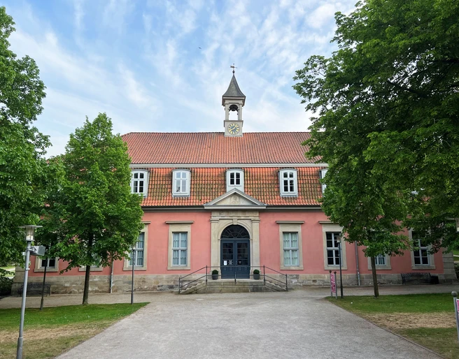 Das Gebäude mit Rosafassade und Glockenturm, umgeben von Bäumen, wirkt ruhig und ansprechend.